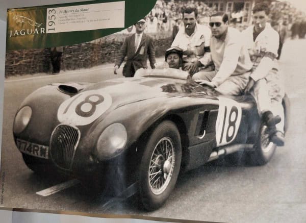 Op een vintage zwart-witfoto uit 1953 is een klassieke raceauto met nummer 18 te zien met drie mannen. Eentje zit op de bestuurdersstoel met een helm en bril, terwijl twee anderen ontspannen op de achterkant zitten. Een Jaguar-embleem voegt een intrigerend detail toe aan deze scène tegen de landelijke achtergrond van Frankrijk.