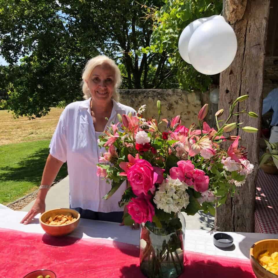 Een persoon met licht haar, gekleed in een wit shirt, staat buiten achter een tafel. De tafel, bedekt met een roestkleurige doek, heeft een vaas met roze en witte bloemen, een schaal met chips en een witte ballon bevestigd aan een houten paal. Weelderig groen omringt de serene omgeving in de omhelzing van de natuur.