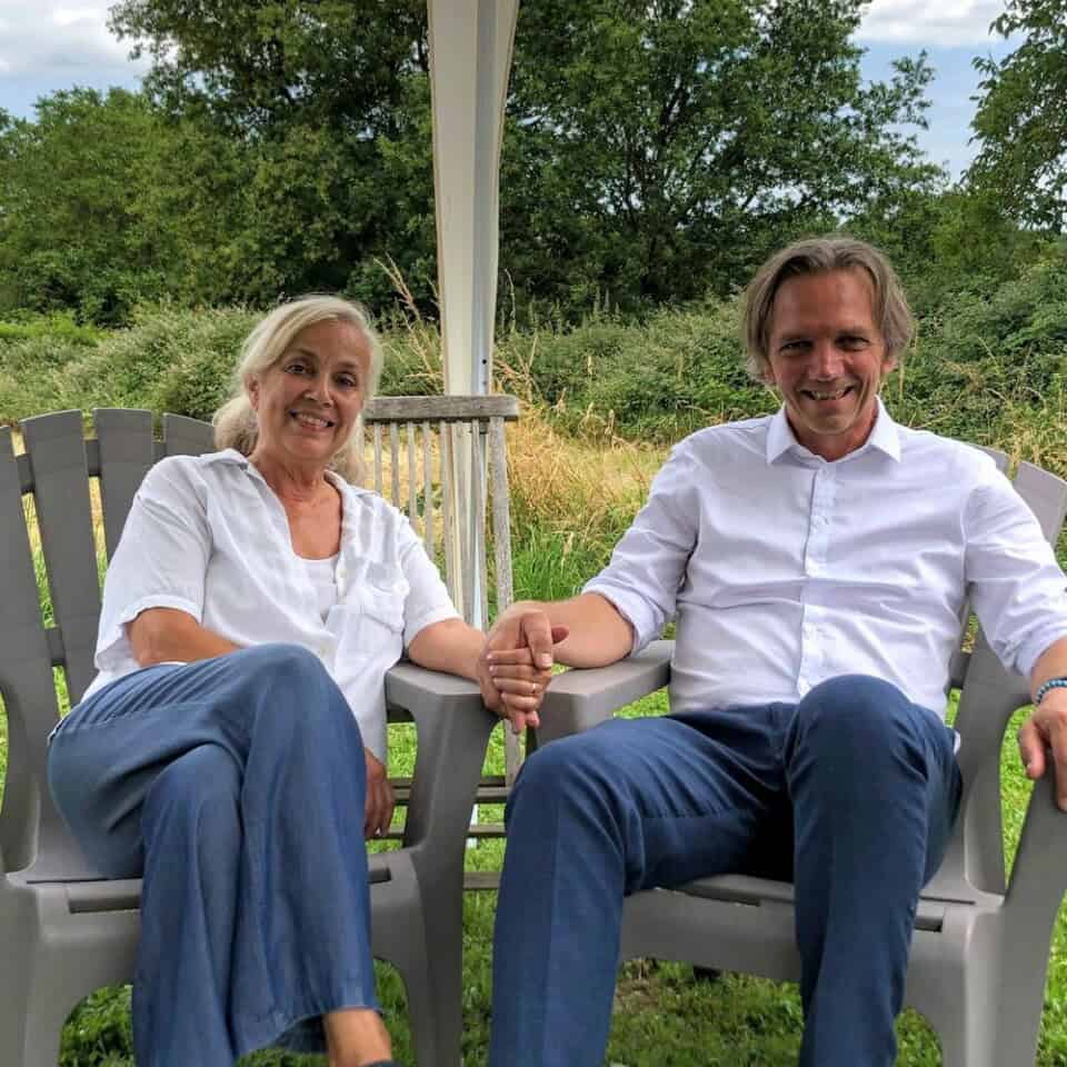 Een lachende vrouw en man zitten buiten onder een luifel op grijze plastic stoelen bij La Camillette gîte. Beiden dragen witte shirts en blauwe broeken. De achtergrond is voorzien van roestkleurige bladeren, wat een vleugje warmte toevoegt aan de serene, natuurlijke omgeving terwijl ze elkaars hand vasthouden, wat duidt op een hechte relatie.