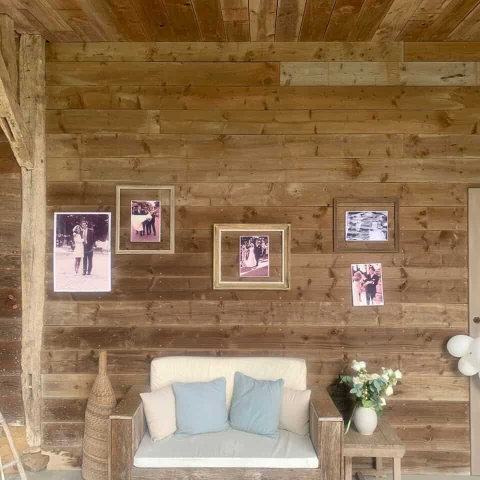 Een gezellige houten kamer in een rustieke gîte heeft een witte bank met blauwgroene en beige kussens tegen een met hout beklede muur. Hierboven zijn vijf ingelijste zwart-witfoto's netjes gerangschikt. Een rieten vaas staat links, terwijl een boeket witte bloemen in een vaas rechts een door de natuur geïnspireerde roestkleur toevoegt.