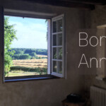 Een rustieke kamer met een groot open raam onthult een schilderachtig uitzicht op velden en bomen onder een blauwe lucht, wat de essentie van het Franse platteland vastlegt. "Bonne Année" staat in witte tekst over de afbeelding heen. In de linkerbenedenhoek staat een logo van een boom en de tekst "Le Camélia - MARQUES DE CAMPAGNE.