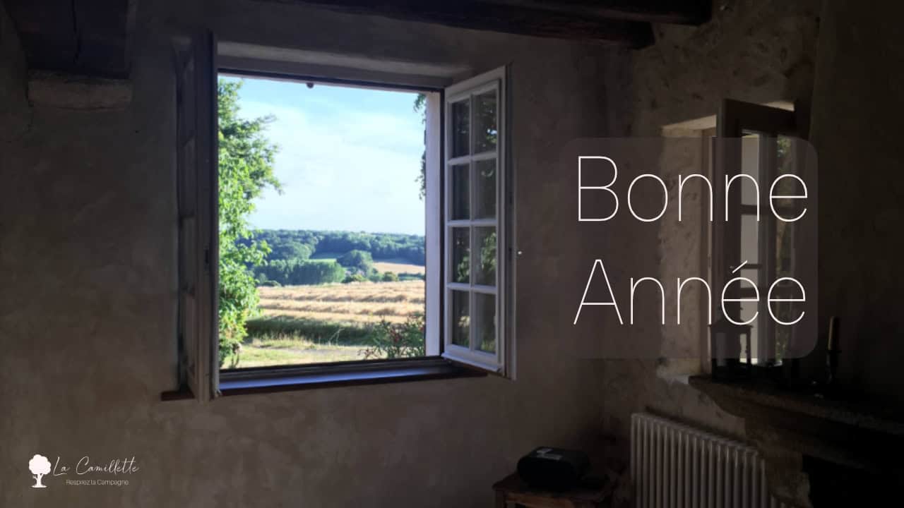 Een rustieke kamer met een groot open raam onthult een schilderachtig uitzicht op velden en bomen onder een blauwe lucht, wat de essentie van het Franse platteland vastlegt. "Bonne Année" staat in witte tekst over de afbeelding heen. In de linkerbenedenhoek staat een logo van een boom en de tekst "Le Camélia - MARQUES DE CAMPAGNE.