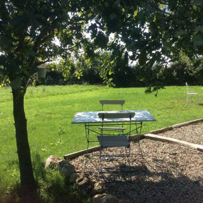 Een metalen tafel en twee stoelen staan onder de schaduw van een boom op een zonnige dag, perfect om te genieten van de natuurlijke schoonheid. De tafel rust op grind, omringd door een grasveld dat doet denken aan een gîte in Frankrijk. Een enkele witte stoel is in de verte zichtbaar te midden van weelderig groen.
