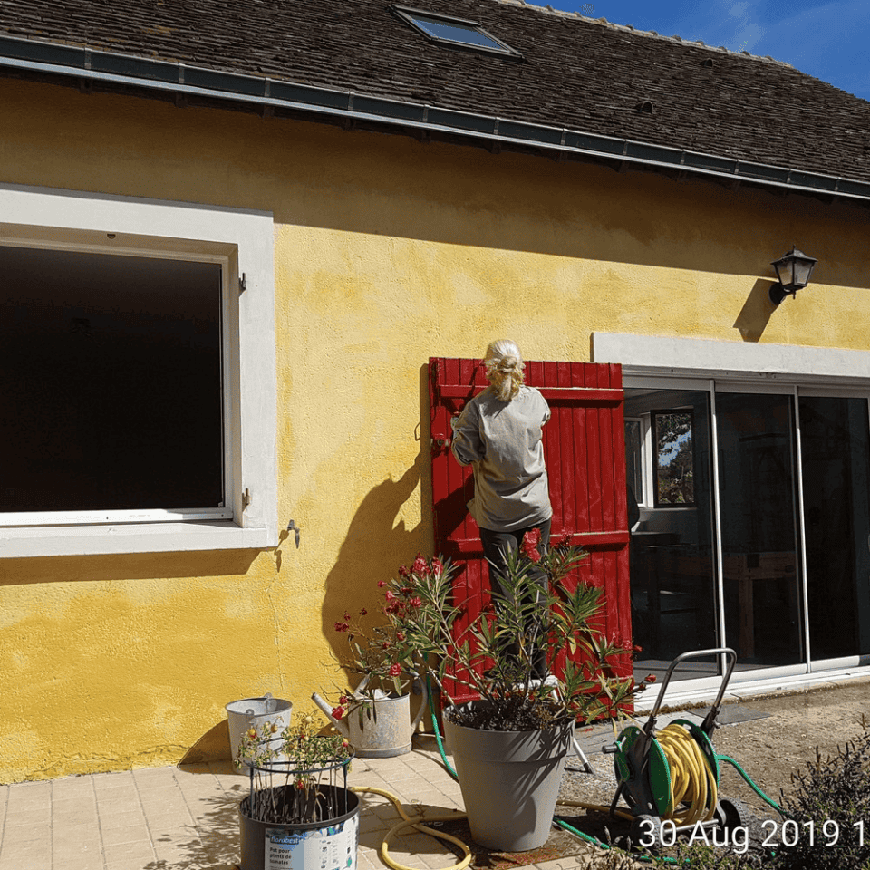 Een persoon met een gezichtsmasker schildert een rood houten luik op een geel huis, wat doet denken aan de charmante gîtes die je op het platteland vindt. In de buurt sieren potplanten en een tuinslang de patio. De heldere, zonnige dag werpt schaduwen op de muur. De datum "30 aug. 2019" is zichtbaar in de rechter benedenhoek.