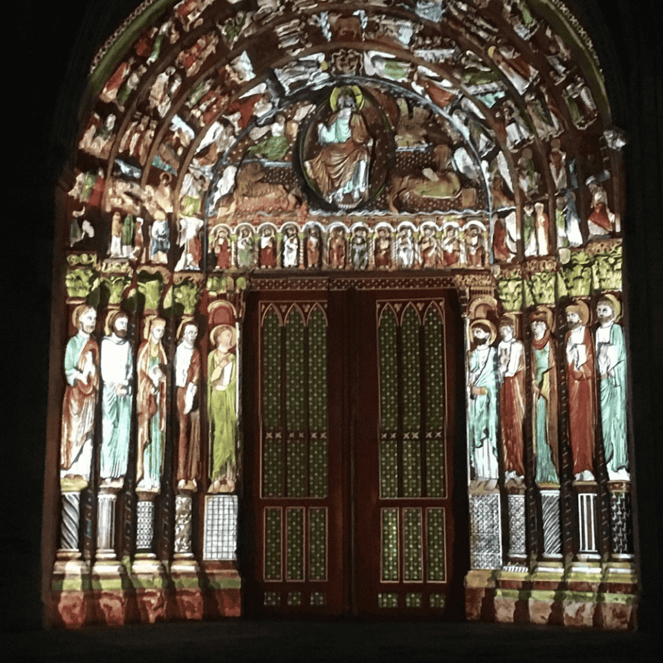De verlichte kathedraalingang in Frankrijk heeft ingewikkelde houtsnijwerken en levendige kleuren. De boog is versierd met religieuze figuren en scènes, badend in licht. Twee grote deuren staan in het midden, omringd door gedetailleerde sculpturen, wat een dramatisch en historisch visueel effect creëert.