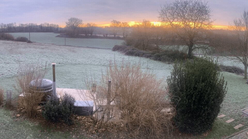 Een ijzig landschap met een lichte laag sneeuw bedekt een veld en bomen onder een pastelkleurige hemel. De zon komt op aan de horizon en werpt een warme gloed. Op de voorgrond is een patio te zien bij gîte La Camillette met een hot tub, omringd door kale struiken en een grote, groenblijvende struik.