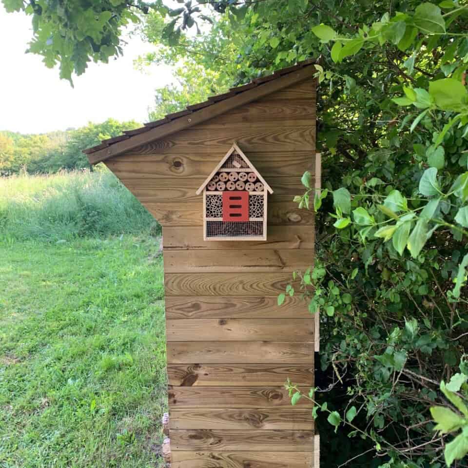 Een houten bijenkorfstructuur staat naast weelderig groen in een omgeving die de charme van Frankrijk oproept. Het heeft een schuin dak en een ingevoegd gebied met houten buizen van verschillende groottes die zijn ontworpen voor bijenhuisvesting, allemaal tegen een achtergrond van levendig gras en dicht gebladerte onder een heldere hemel.