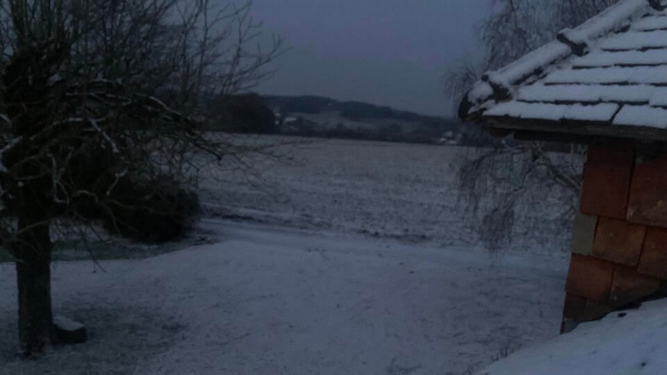 Een besneeuwd landschap bij zonsopgang onthult een met sneeuw bedekt veld en een paar bomen aan de linkerkant. Het dak van "La Camillette", met zijn roestkleurige, met sneeuw bestoven dakspanen, is zichtbaar aan de rechterkant. Op de achtergrond rusten een klein bos en heuvels onder een bleke, vroege ochtendhemel.