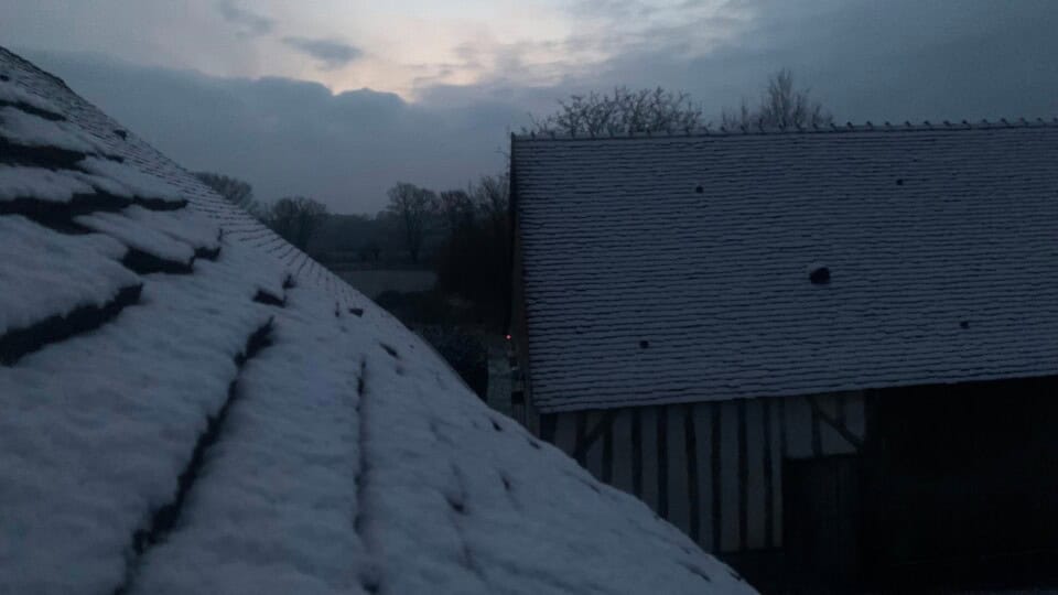 Daken bedekt met een dun laagje sneeuw in de schemering creëren een sereen plattelandsgezicht. De lucht is bewolkt met een flauwe gloed van de ondergaande zon. Bomen zijn silhouetten op de achtergrond en het zwakke licht suggereert een koude, vroege winteravond - de perfecte achtergrond voor een rustieke gîte.