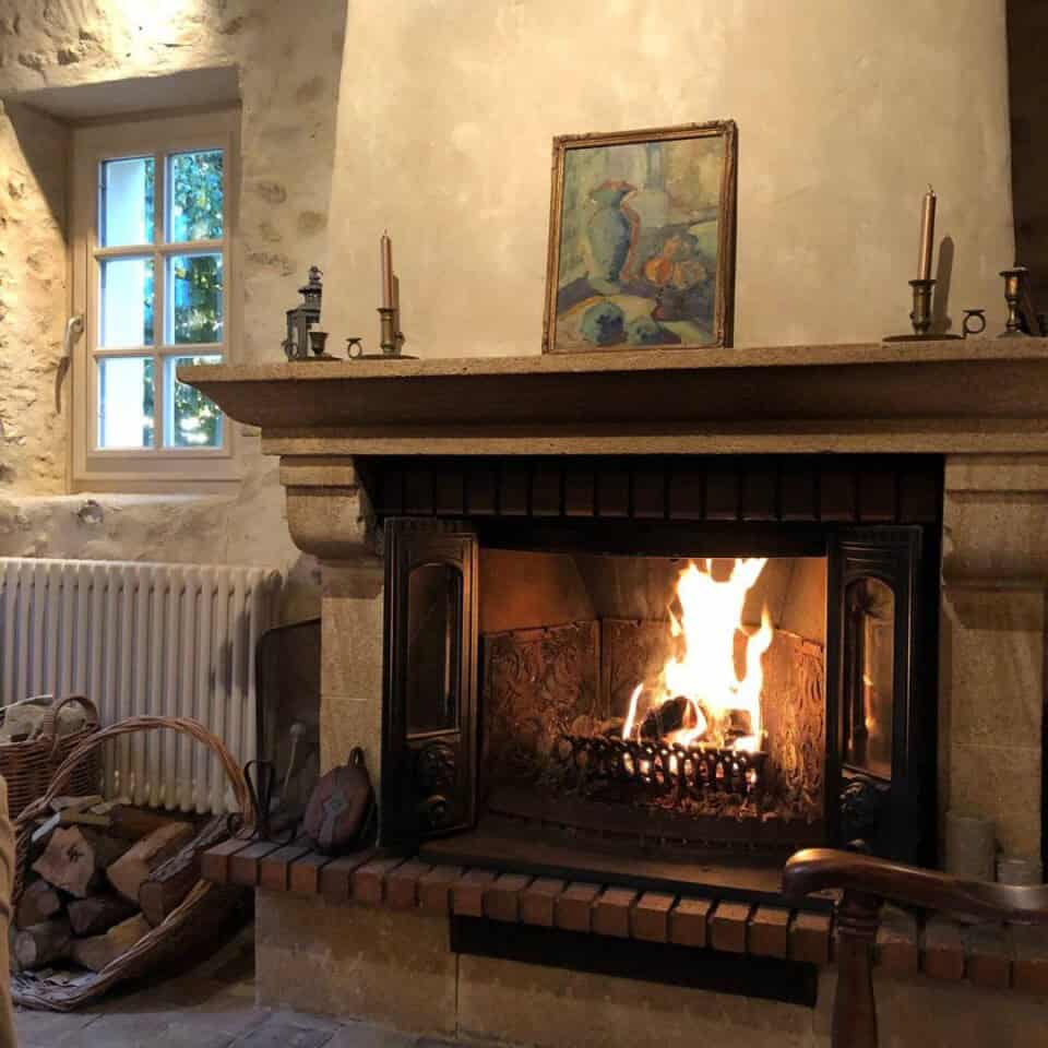 Een gezellige stenen open haard met een brullend vuur bepaalt de sfeer in deze rustieke gîte. Een schilderij, kandelaars en kleine decoratieve items sieren de schouw. Een gevlochten mand met brandhout leunt tegen de muur terwijl zacht, natuurlijk licht door een wit omrand raam filtert, wat een sfeer van roest creëert die doet denken aan Frankrijk.