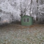 Een klein groen schuurtje met ramen met witte omlijsting staat in een met vorst bedekt bos. De omringende bomen zijn bedekt met zware vorst, wat een pittoresk plattelandsgezicht creëert. De grond is licht bestrooid met vorst en gevallen bladeren, wat bijdraagt aan de koude sfeer van deze natuurlijke omgeving.