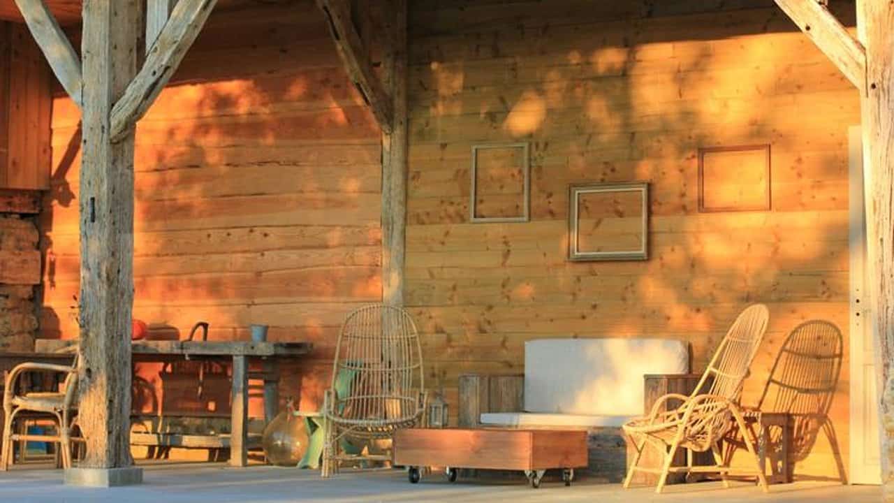 Een zonnig houten terras met rustiek meubilair, waaronder rieten stoelen en een bank met een houten salontafel. Lege fotolijsten sieren de muur terwijl schaduwen van nabijgelegen bomen patronen vormen, waardoor een gezellige plattelandssfeer ontstaat vol natuur en roest.