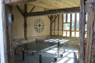 Een zonnige kamer met zichtbare houten balken en rustieke muren heeft een pingpongtafel. Een grote, decoratieve klok hangt aan de muur. Zonlicht stroomt door hoge ramen en werpt schaduwen op de vloer. De gîte biedt een pastoraal uitzicht dat zichtbaar is door de ramen.