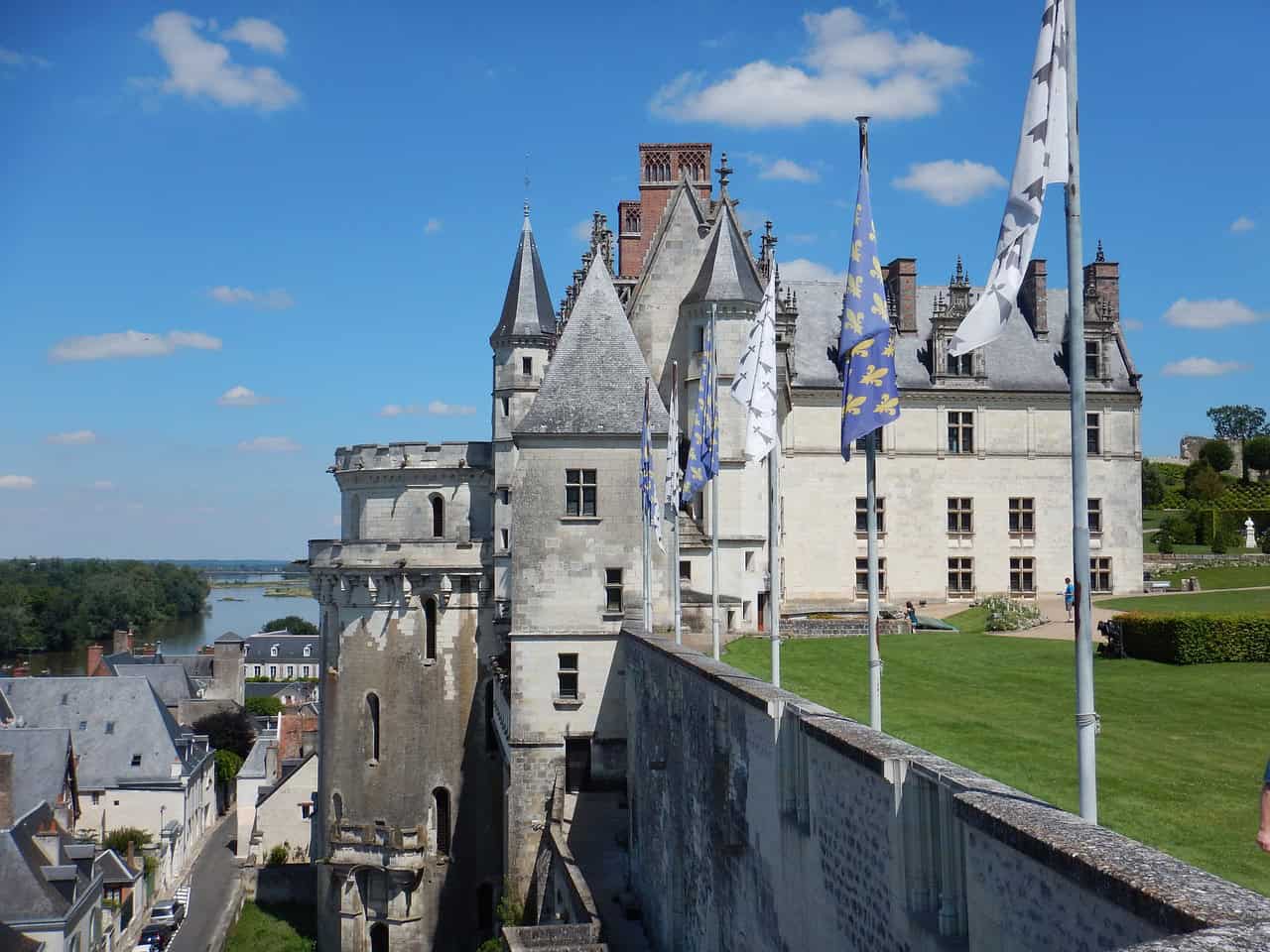 Een historisch stenen kasteel met puntige torens en schoorstenen steekt trots af tegen een helderblauwe lucht. Het kasteel wordt omringd door een verzorgd gazon, verschillende vlaggen op palen en het vredige platteland. Op de achtergrond stroomt een rivier zachtjes langs aangrenzende daken van kleinere gebouwen, terwijl donzige wolken de lucht bedekken.