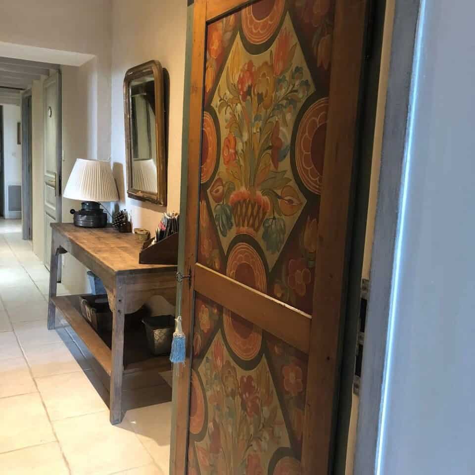 Een gang in La Camillette in Frankrijk heeft een vintage houten deur met kleurrijke, sierlijke bloemenpatronen. Een houten consoletafel met een decoratieve lamp, spiegel en kleine spullen staat tegen de muur. De vloer is betegeld in lichtbeige en zacht natuurlijk licht vult de uitnodigende gîteruimte.