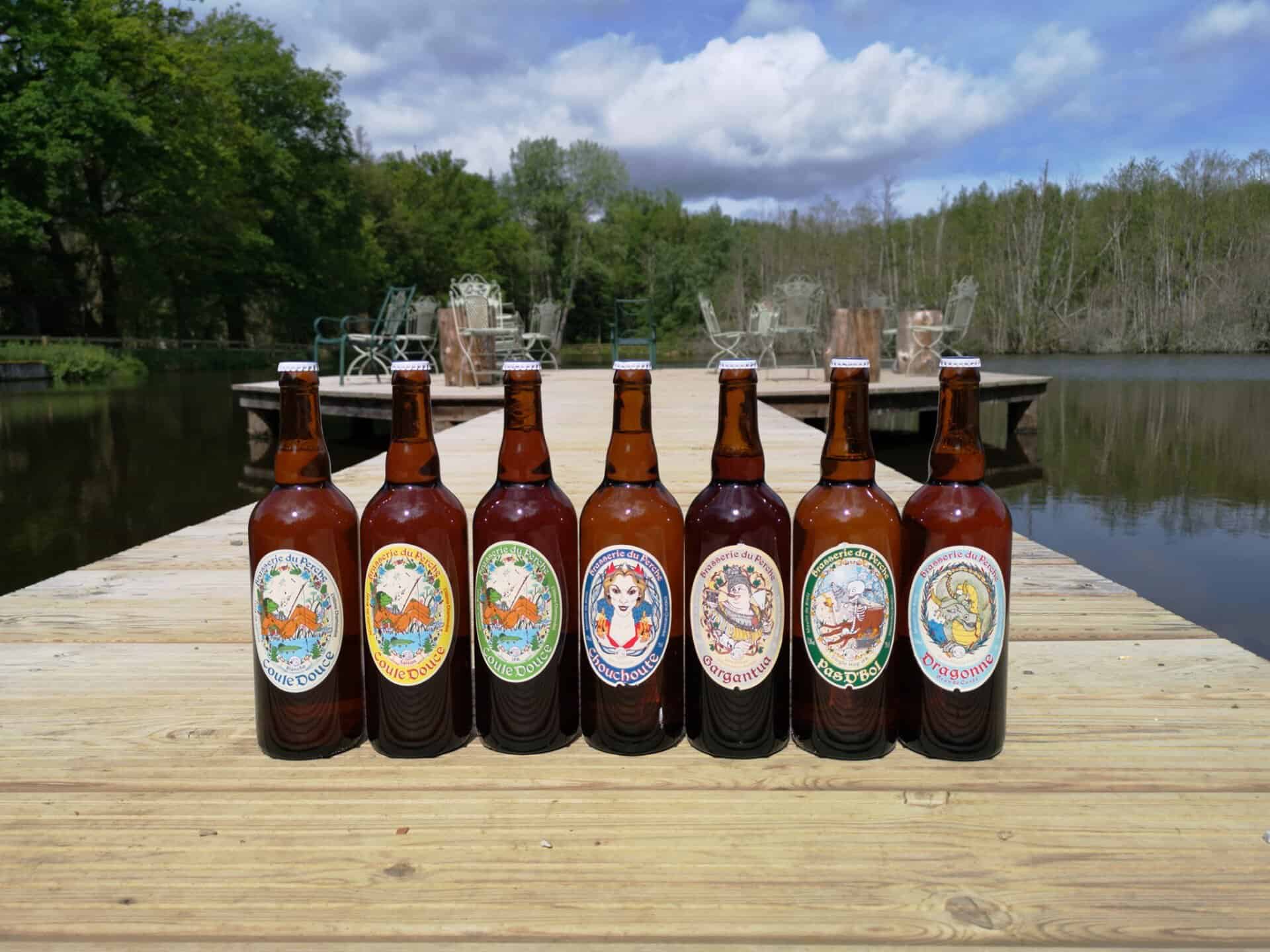 Acht bruine bierflessen met kleurrijke etiketten staan opgesteld op een houten steiger die zich uitstrekt over een kalm meer, wat doet denken aan een gezellig vakantiehuisje. Bomen grenzen aan het meer en ligstoelen zijn zichtbaar op de achtergrond onder een gedeeltelijk bewolkte lucht, wat een ontspannen sfeer creëert.