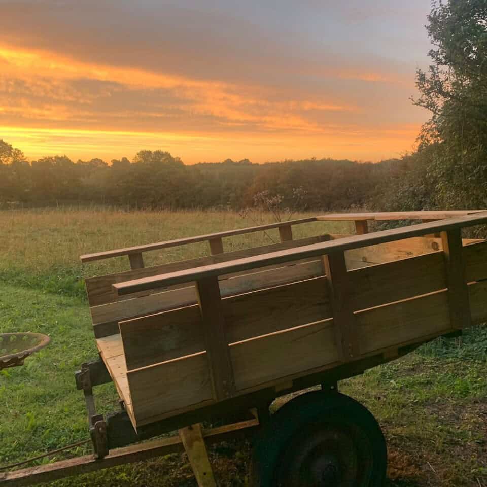 Een houten kar met een rustiek ontwerp staat op een grasveld bij zonsondergang in het hart van het Franse platteland. De lucht straalt levendige oranje en gele tinten uit, die een warme gloed werpen over deze charmante gîte-setting. Bomen en struiken volgen de horizon terwijl de schaduw van de kar over het gras danst.