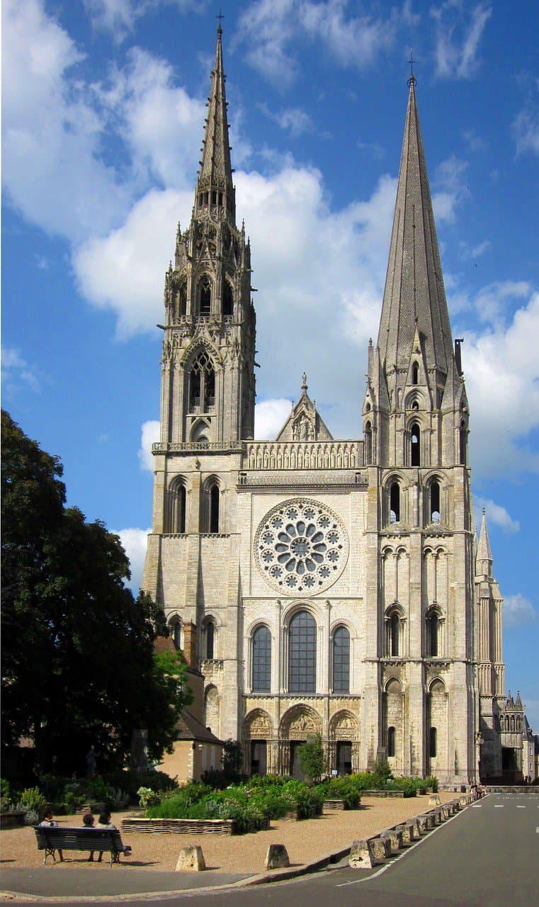 De afbeelding toont het vooraanzicht van de kathedraal van Chartres in Frankrijk, met twee asymmetrische torenspitsen tegen een blauwe lucht met wolken. Het bevat een groot roosvenster, sierlijk steenwerk en gedetailleerde sculpturen. Een boom staat links naast een bank langs het pad, met stenen barrières langs het gebied.