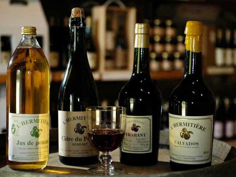 Een selectie van vier flessen op een houten vat in een schemerige omgeving, perfect om van te genieten in een knusse gîte. Elke fles heeft een eigen etiket, waaronder cider en calvados. Voor de flessen staat een glas vol met donkere vloeistof, terwijl planken met meer flessen zachtjes vervagen op de achtergrond.