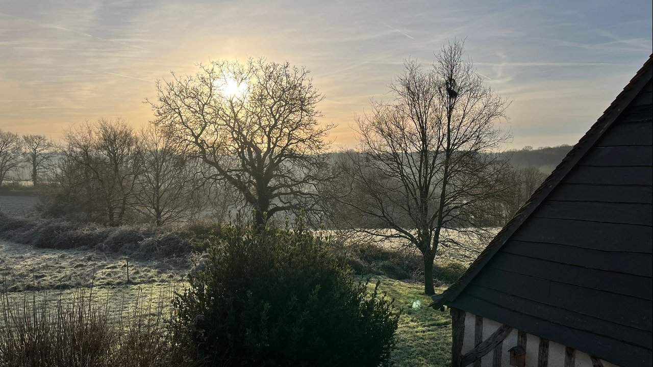 Zonsopgang boven een ijzig plattelandslandschap met kale bomen die lange schaduwen werpen op een met gras bedekt veld. De zon, gedeeltelijk verduisterd door takken, creëert een warme gloed in de lucht. Een gebouw met een steil, donker dak is zichtbaar op de voorgrond, wat de serene en koude natuursfeer van Frankrijk oproept.