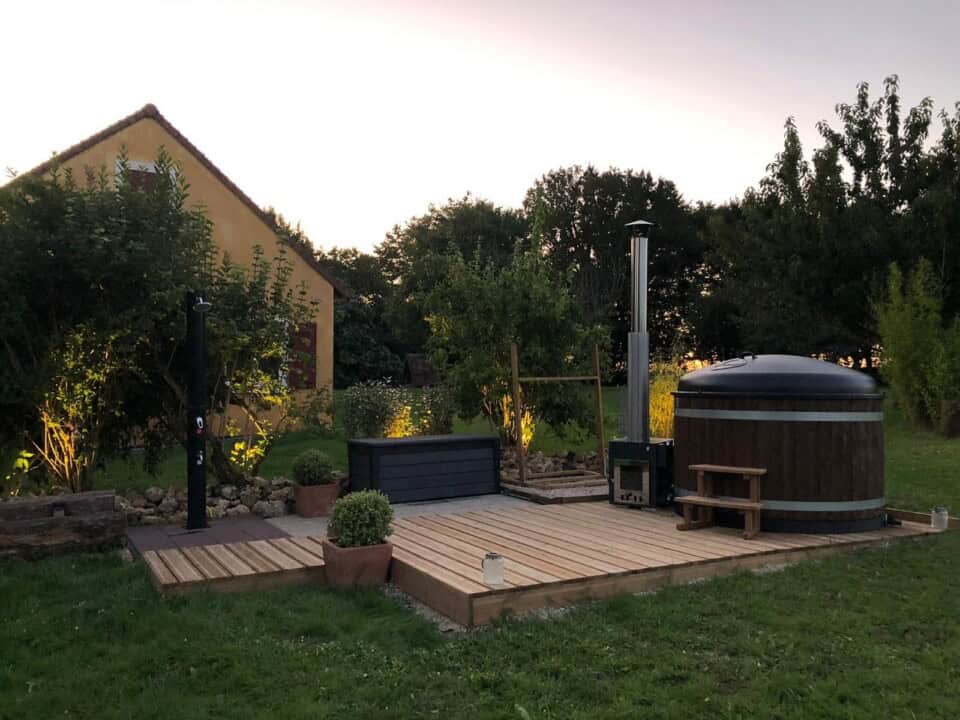 Een rustige tuin bij zonsondergang met een houten terras met potplanten en een klein bankje. Een charmant huis en weelderig groen omringen het gebied, met zachte verlichting die de planten verlicht. Aan de rechterkant draagt een houten hottub met een schoorsteen bij aan deze uitnodigende buitenruimte, die een vleugje schoonheid van de natuur biedt.