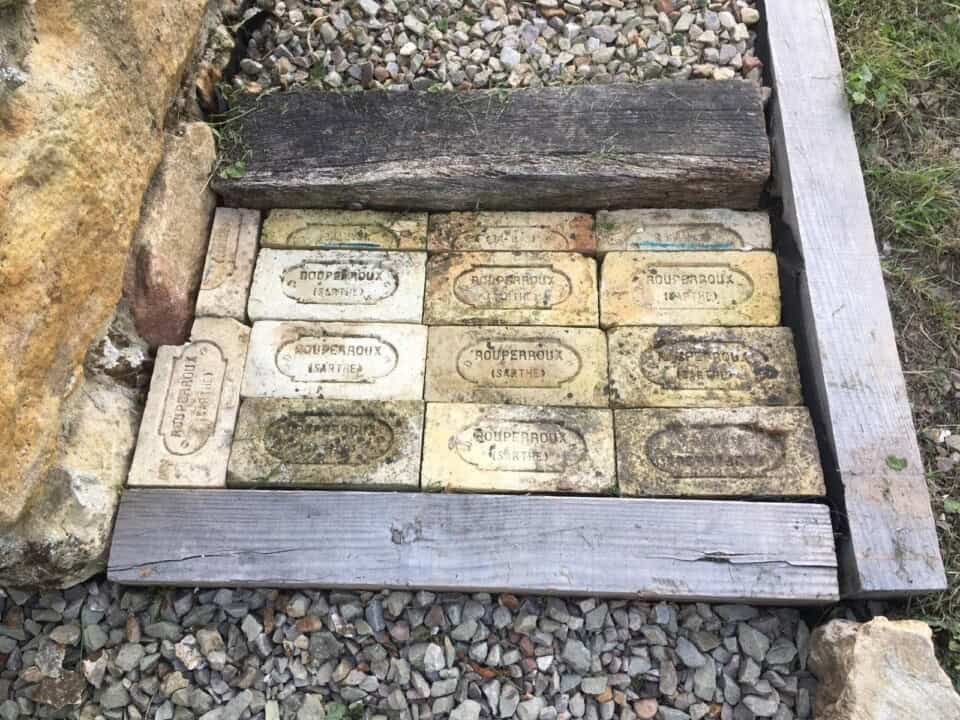 Een pad van rechthoekige bakstenen met het label "ROUFBROUX SAMBRE" wordt omlijst door houten balken en omzoomd met kleine stenen. Een grotere ruwe steen markeert de linkerbovenhoek en gras piept van rechts. Deze rustieke omgeving brengt een vleugje natuur dat doet denken aan Frankrijk.