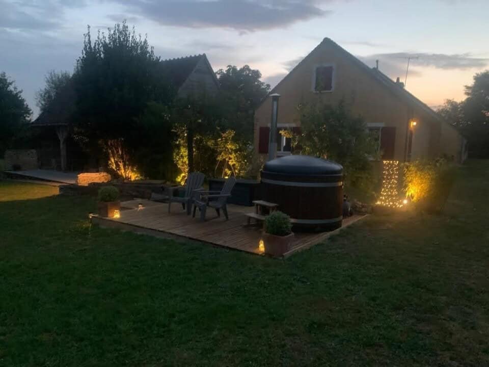 Een gezellig buitentafereel bij schemering onthult een houten terras met twee Adirondack stoelen en een ronde houten hottub. Zachte verlichting verlicht bomen en een huis met gele muren en rode luiken, wat doet denken aan de charmante stijl die vaak in Frankrijk te vinden is. De tuin heeft gras en subtiele sfeerverlichting.