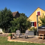 Een charmante gîte met een gele gevel en rode luiken staat in een weelderige tuin. Een houten terras heeft Adirondack-stoelen en een ronde hot tub. Bomen en struiken omringen het gebied, terwijl een helderblauwe lucht het rustige, zonnige tafereel completeert dat doet denken aan het landelijke Frankrijk.
