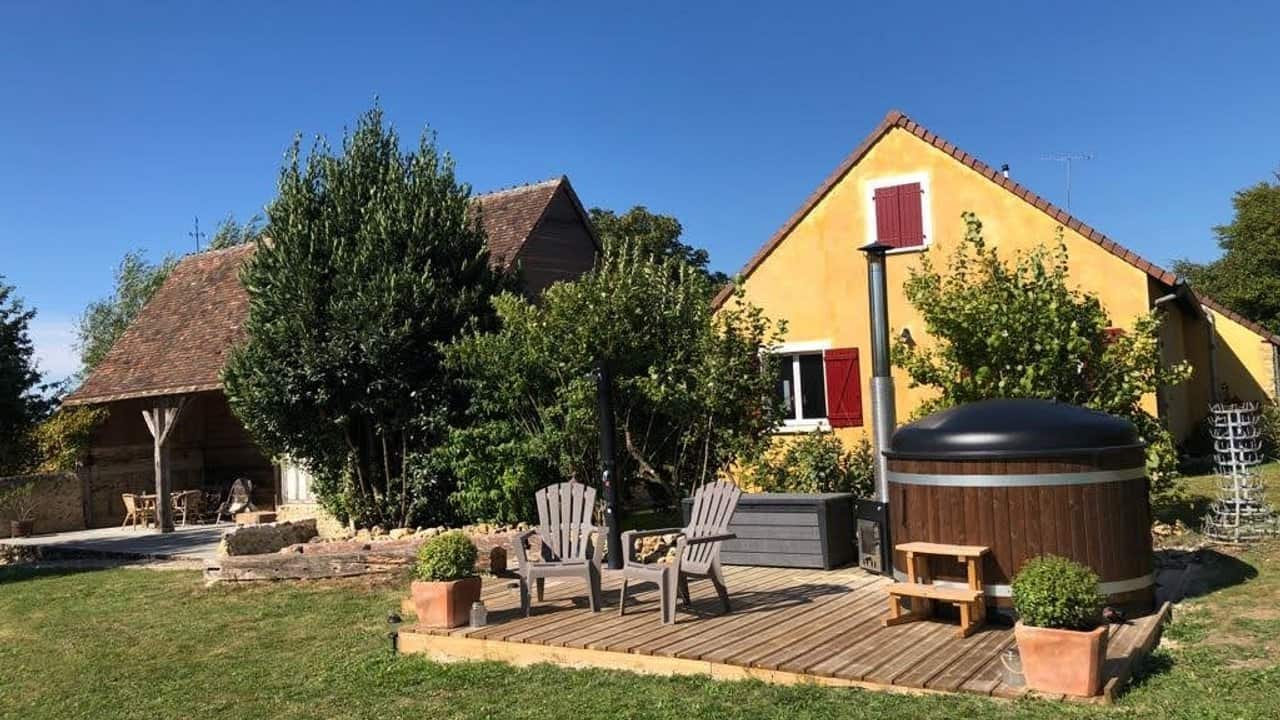 Een charmante gîte met een gele gevel en rode luiken staat in een weelderige tuin. Een houten terras heeft Adirondack-stoelen en een ronde hot tub. Bomen en struiken omringen het gebied, terwijl een helderblauwe lucht het rustige, zonnige tafereel completeert dat doet denken aan het landelijke Frankrijk.