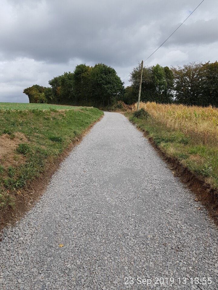 Een grindpad slingert door het platteland, begrensd door gras en een maïsveld aan de rechterkant. Het pad leidt naar een cluster van dichte, bladrijke bomen onder een bewolkte lucht. Een houten paal met een draad is in de buurt zichtbaar. In de hoek staat in vette letters "23 sep 2019, 13:13:55".