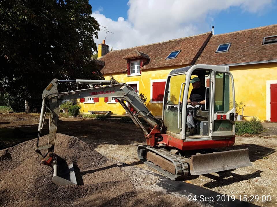 Een persoon bedient een rood-witte Takeuchi minigraafmachine en verplaatst aarde voor een charmante gele gîte met rode luiken en dak. De tuin heeft stukken gras en grind, met een gedeeltelijk bewolkte lucht erboven. De datumstempel luidt "24 Sep 2019 15:29:00.