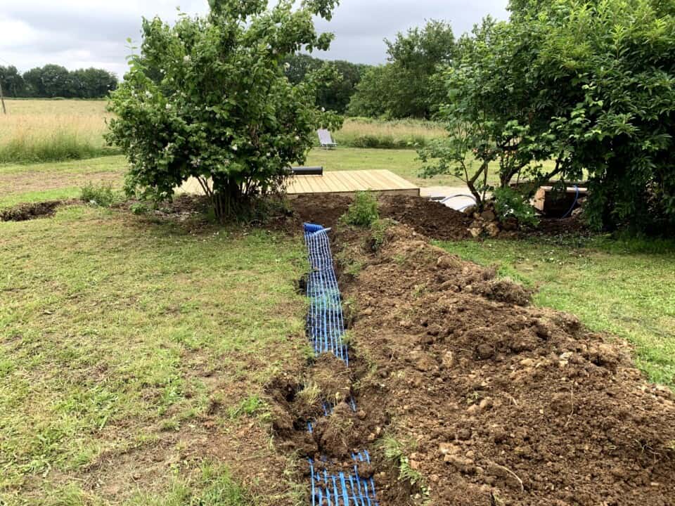 Een gegraven sleuf in een grasveld heeft blauwe buizen blootgelegd, omzoomd door een hoop aarde. Op de achtergrond omlijsten weelderige groene struiken een nieuw houten dek en steken bomen af tegen een bewolkte lucht. Het is een doorlopend landschapsproject dat typerend is voor het Franse platteland, misschien in de buurt van een knusse gîte.