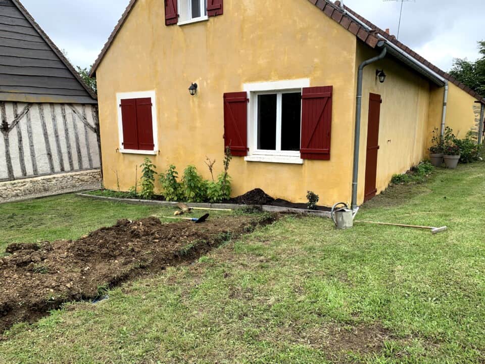 Een klein geel huis met rode luiken staat in een tuin op het vredige platteland. Vers bewerkte grond vormt een greppel over het gazon op de voorgrond, met een schop en gieter in de buurt. Een wit raam en een klein portieklicht zijn zichtbaar, omgeven door groen gras en een paar planten.