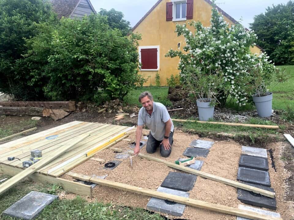 Een man knielt op de grond en werkt op een houten terras in een tuin. Hij houdt een gereedschap vast, omringd door planken en stenen. Achter hem staat een geel huisje met rode luiken en weelderig groen, als bloeiende struiken in grote potten. Dit buitentafereel ontvouwt zich onder een bewolkte lucht, en belichaamt echte roest - perfect voor degenen die de rustige omhelzing van de natuur zoeken in de gîte.