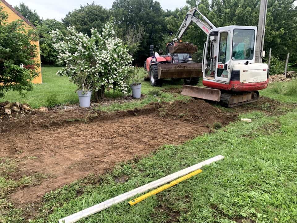 Een minigraafmachine egaliseert een stukje aarde in een grasveld, perfect voor een rustieke gîte-setting. In de buurt staan twee bloeiende struiken in potten. Een geel gebouw is gedeeltelijk zichtbaar op de achtergrond met een hark en een lange vlakte op het gras, alles omgeven door groen en bomen.