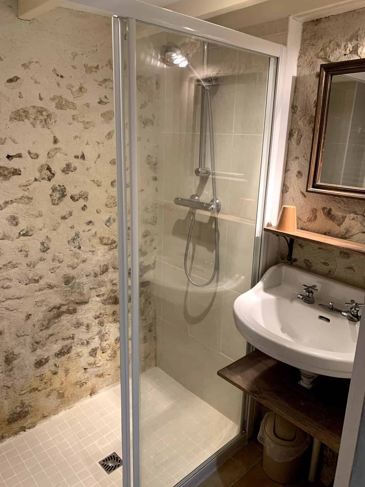 Een rustieke badkamer met een stenen muur, met een glazen douche met een chromen douchekop. Ernaast staat een witte keramische wasbak op een houten plank, die doet denken aan het Franse platteland. Boven de wasbak hangt een rechthoekige spiegel met een bruine lijst en een kleine lamp op de plank.
