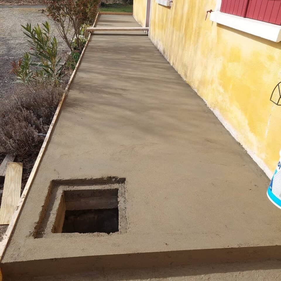 Een vers gestort betonnen pad loopt langs een geel huis met rode luiken, genesteld in het landschap. Het pad heeft een vierkant gat aan één kant, misschien voor toegang tot nutsvoorzieningen of beplanting. Houten planken omzomen het pad en weelderig gebladerte omzoomt de buitenste rand onder een heldere en zonnige hemel.