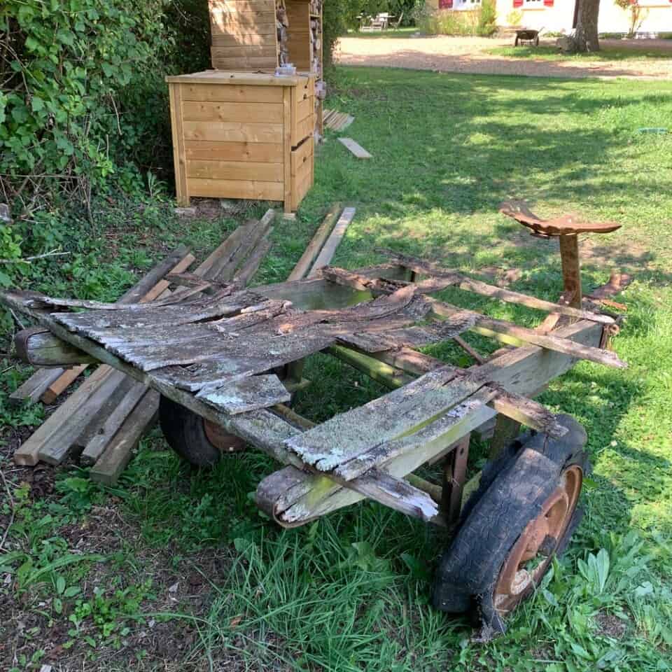 Een houten kar met schilferende planken staat op grasachtige grond, de grote wielen zijn leeg. In de buurt is een composthoop zichtbaar, omringd door verspreide stukken hout. Op de achtergrond zijn bladrijke struiken en een boom te zien, wat doet denken aan een tuinomgeving in de buitenlucht die doet denken aan La Camillette in Frankrijk, een perfect toevluchtsoord.