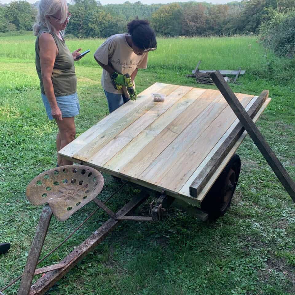 Twee mensen werken aan een houten constructie op een trailer in een grasveld. De een, gekleed in een tanktop en korte broek, observeert terwijl de ander, gekleed in een T-shirt en jeans, een boormachine gebruikt op de planken. Een oude metalen stoel is bevestigd aan het frame, omringd door hints van de natuur en bomen op de achtergrond.