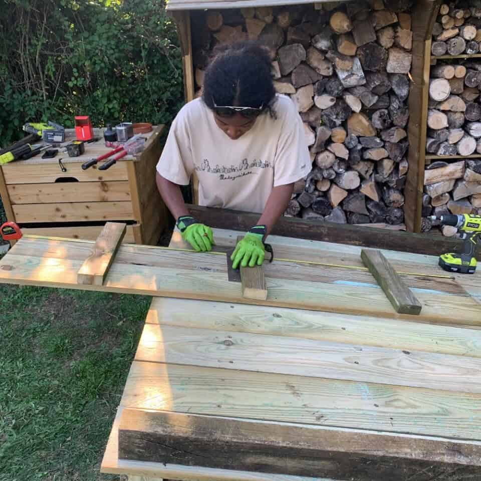 In een grasveld omzoomd door bomen, zet een persoon in een wit T-shirt en groene handschoenen een houten constructie in elkaar. Ze werken met gereedschap op een tafel bij een houtstapel, omringd door spullen zoals klemmen en een boor. De setting roept de rustieke charme van het platteland op.