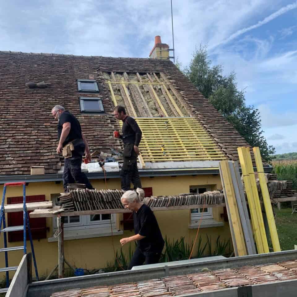 Drie bouwvakkers renoveren een dak. Twee staan op een houten platform dat wordt ondersteund door ladders, terwijl een ander op de grond staat. Het huis, dat doet denken aan de huizen op het Franse platteland, is geel met witte kozijnen. De heldere lucht erboven toont ijle wolken die voorbij drijven.