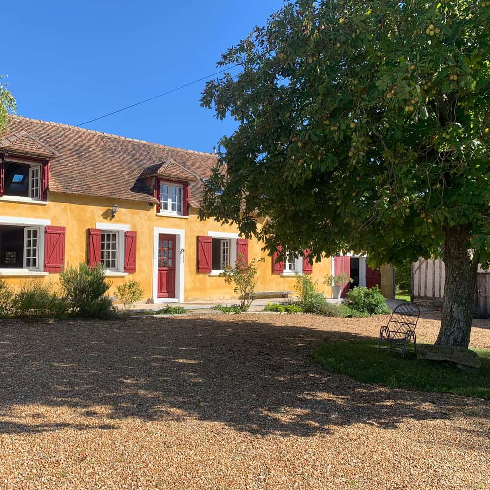 Een charmant huis met twee verdiepingen, La Camillette, met gele pleistermuren en rode luiken staat onder een helderblauwe lucht in het platteland. Een grote bladerrijke boom biedt schaduw over een grindbinnenplaats aan de voorkant. Een metalen bank staat onder de boom. Het huis heeft een rustiek pannendak en meerdere ramen.