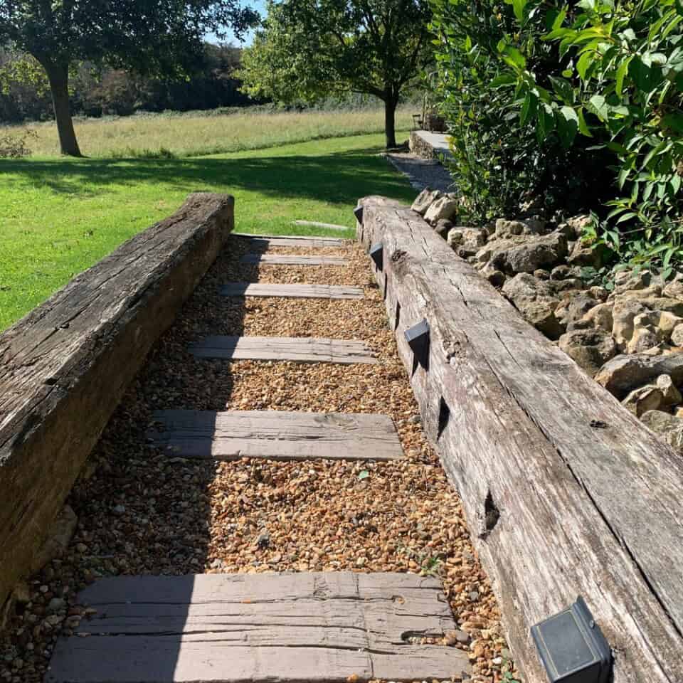Een grindpad met houten treden, begrensd door grote houten balken, slingert zich een weg door rotsen en dicht groen gebladerte. Aan de rechterkant leidt het je zachtjes naar een open grasveld met bomen onder een helderblauwe lucht, wat een waar gevoel van roest en natuurlijke schoonheid oproept.