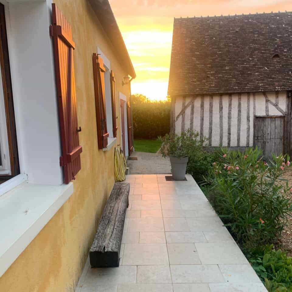 Een charmant huisje met gele muren en rode luiken wordt verlicht door de ondergaande zon. Een houten bank staat op een betegeld pad dat leidt naar een rustiek houten gebouw, omringd door het vredige platteland. Groene planten omzomen het pad en de lucht gloeit met warme zonsondergangtinten.
