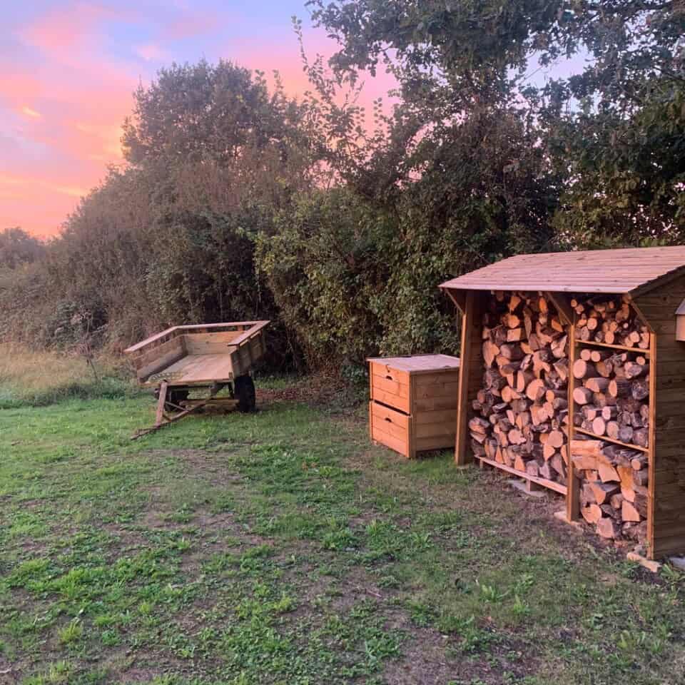 Een vredige buitenomgeving met een houten schuur vol boomstammen. In de buurt staan een kleine houten kist en een geparkeerde tweewielerkar op het grasveld. Dichte bomen en struiken vormen de achtergrond onder een roze en oranje zonsonderganghemel, wat een ideale achtergrond creëert voor een rustieke gîte-ervaring.