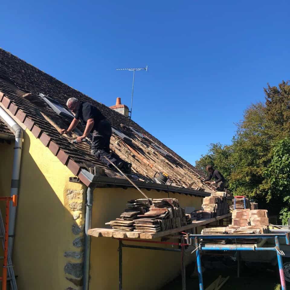 Twee arbeiders repareren een schuin dak van een geel huis onder een helderblauwe lucht. Ze hanteren houten tegels, waarvan sommige op steigers staan. Een schoorsteen en antenne zijn te zien en bomen sieren de achtergrond. Het doet denken aan het dagelijks leven op het charmante platteland van Frankrijk.