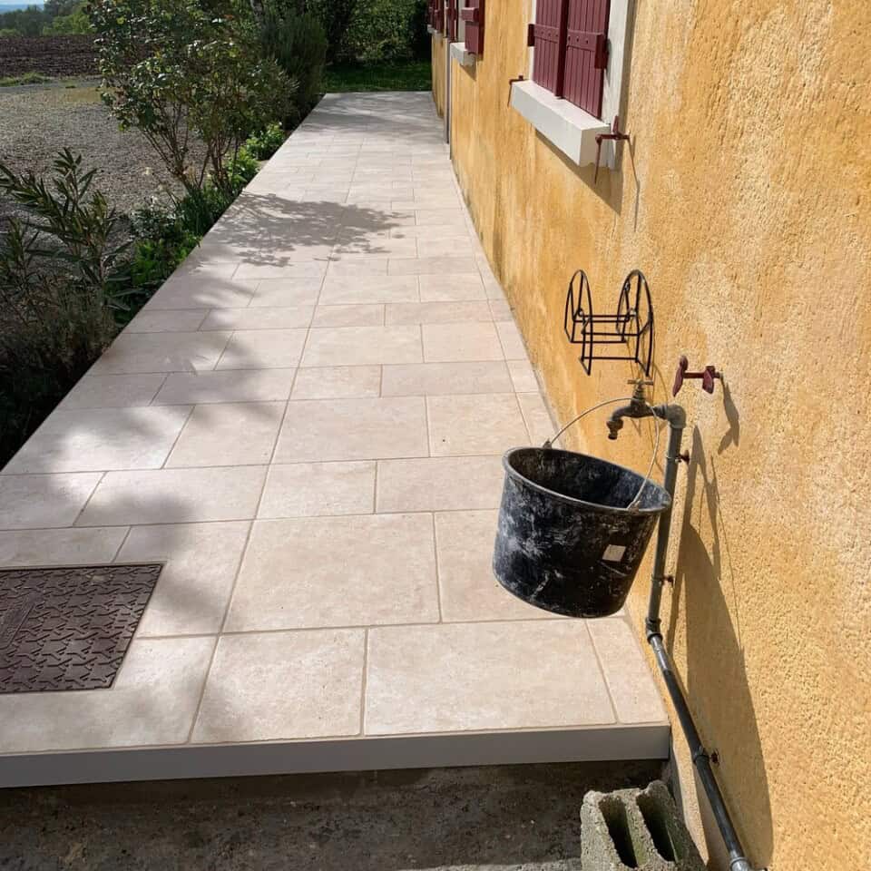 Een betegelde loopbrug langs een gele stucwerkmuur met rode luiken toont een zwarte emmer die aan een buitenkraan hangt. Het pad, omzoomd door een tuin vol levendige planten, leidt naar de ingang van de gîte. Een bruine deurmat ligt bij de deur terwijl zonlicht schaduwen op het tafereel werpt.