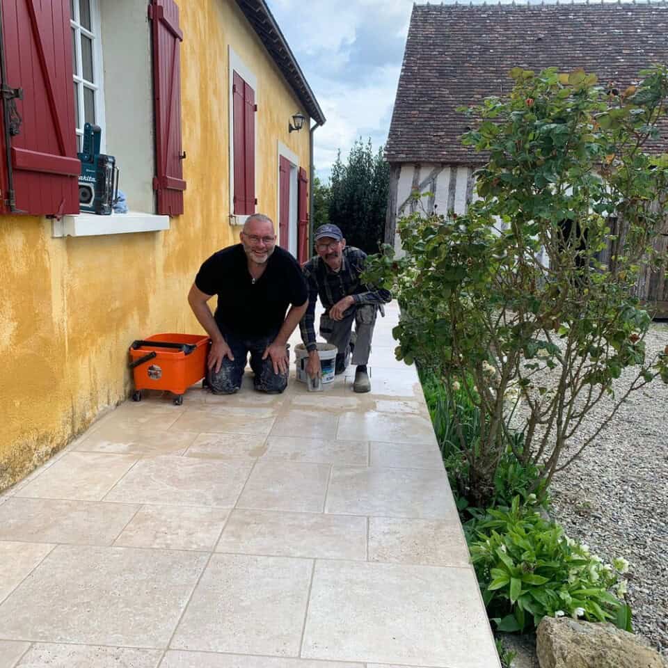 Twee mannen knielen op een nieuw aangelegde stenen patio naast een geel huis met rode luiken, lachend en met gereedschap in de hand. De een draagt een zwart shirt, de ander een geruit shirt en een pet. Er staat een oranje gereedschapskist in de buurt. Een potplant voegt charme toe, terwijl op de achtergrond een schilderachtig houten gebouw hint naar het platteland van Frankrijk.