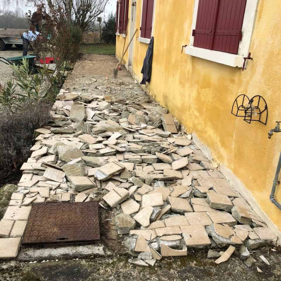 Een beschadigd buitenpad met verspreide, gebroken tegels ligt naast een geel gebouw. De tegels, ooit gerangschikt in een patroon, liggen nu in wanorde, met enkele ontbrekende stukken. Twee rode luiken voor ramen zijn zichtbaar boven het pad, en een metalen wanddecoratie is in de buurt gemonteerd.