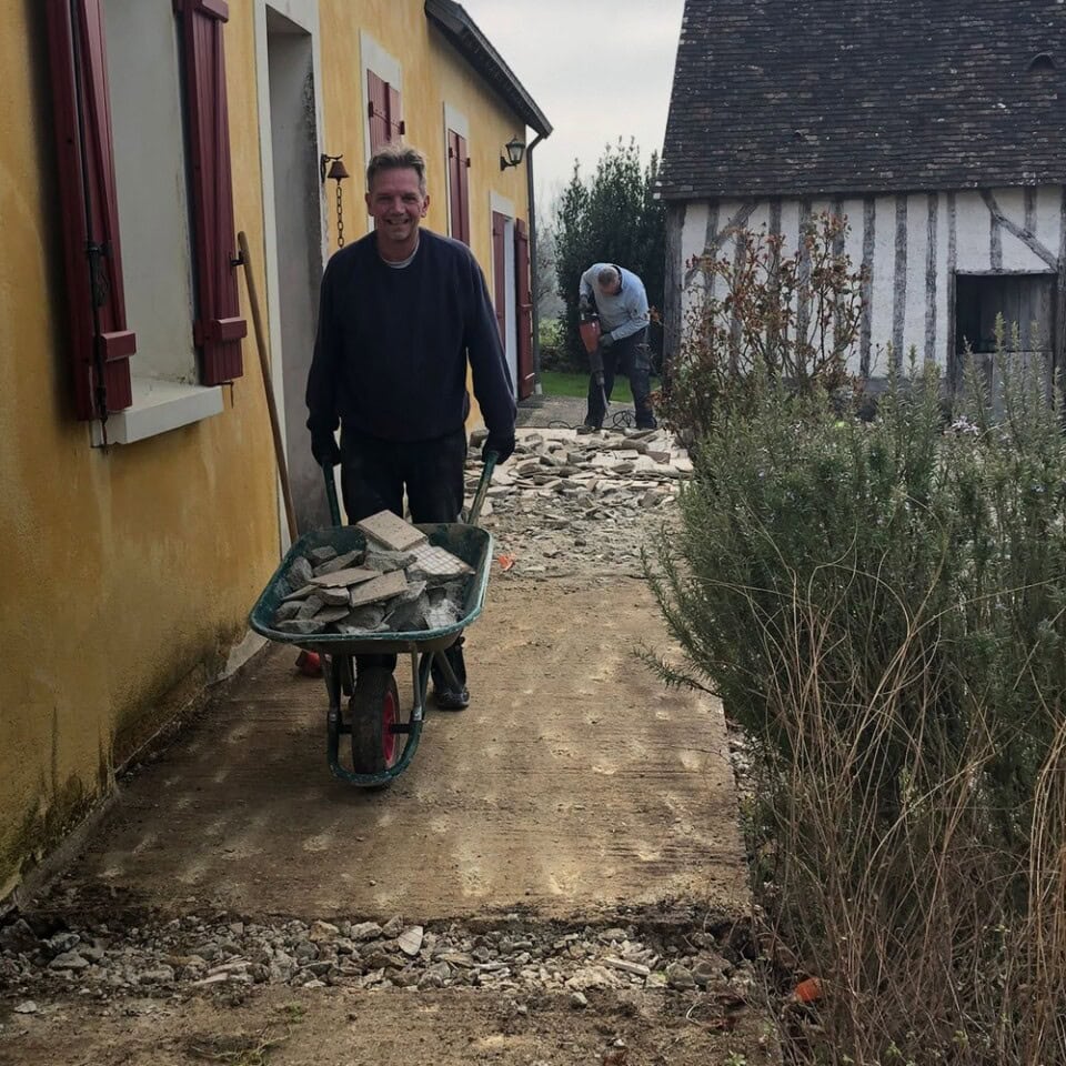 Een man duwt een kruiwagen vol met puin over een pad naast een geel gebouw met rode luiken. Een ander persoon werkt met puin op de achtergrond. In de buurt staat een klein wit vakwerkgebouw terwijl planten het pad omzomen, wat een natuurlijk element aan de scène toevoegt.